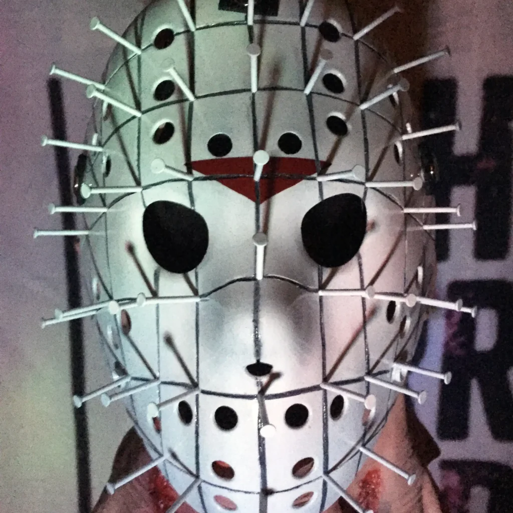 Hand-painted Hellraiser Pinhead Hockey Mask with metallic pins, white face, and black eyes on white ABS plastic, held against a dark background.