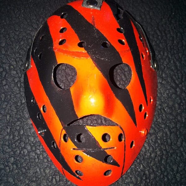 Close-up of the WWE Kane mask featuring bold red and black designs, with removable elastic straps against a textured black background.